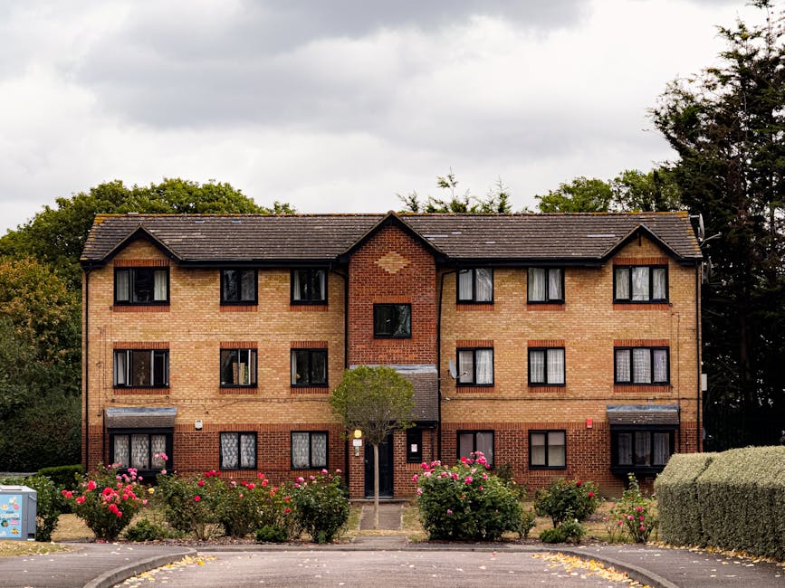 The image depicts a three-story brick residential building with multiple windows, including some with black frames and window boxes. In front of the building, there is a well-maintained garden area fe